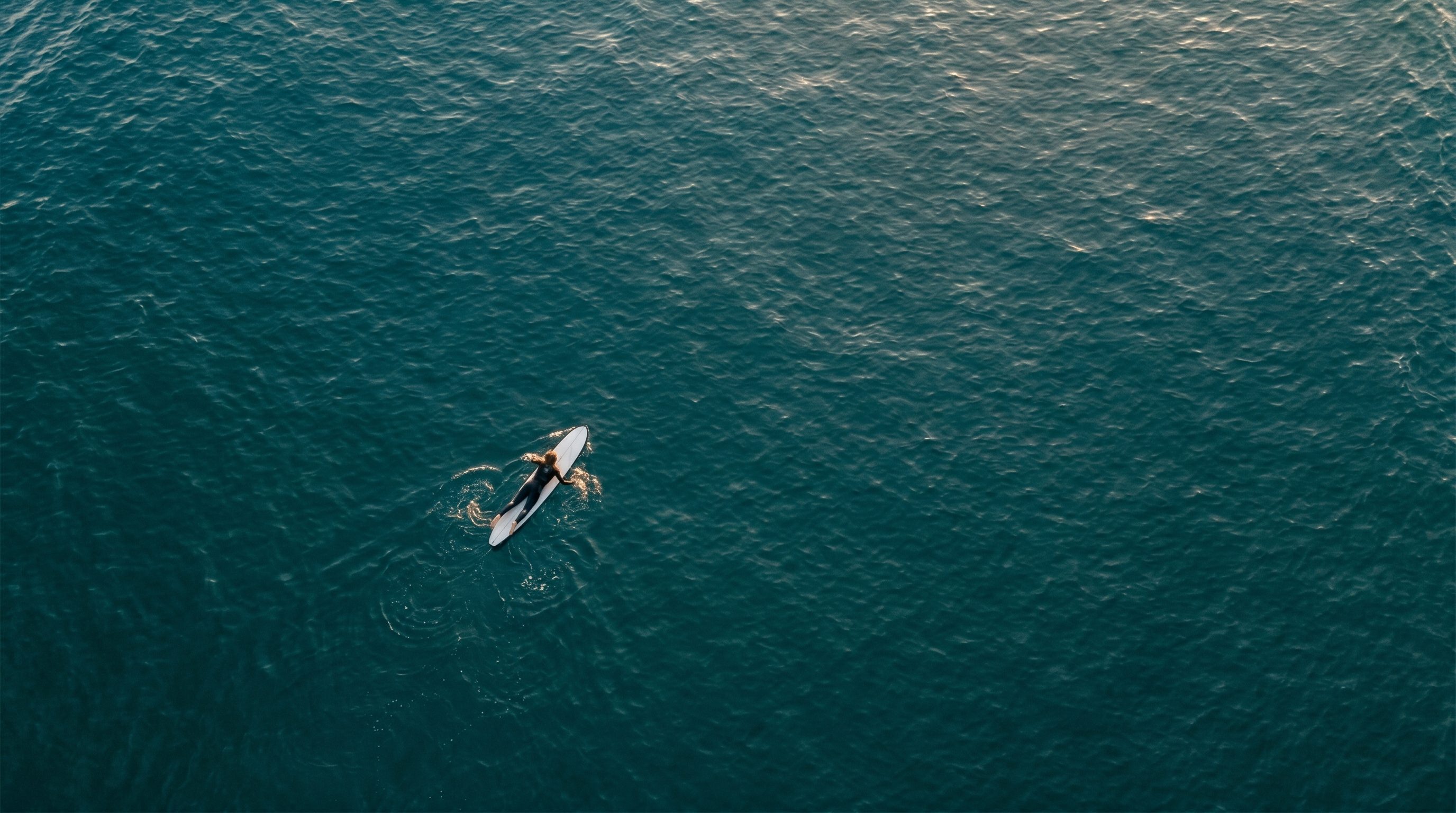 Vue aérienne d'un surfeur seul ramant sur sa planche au milieu d'un océan profond, lumière dorée du matin